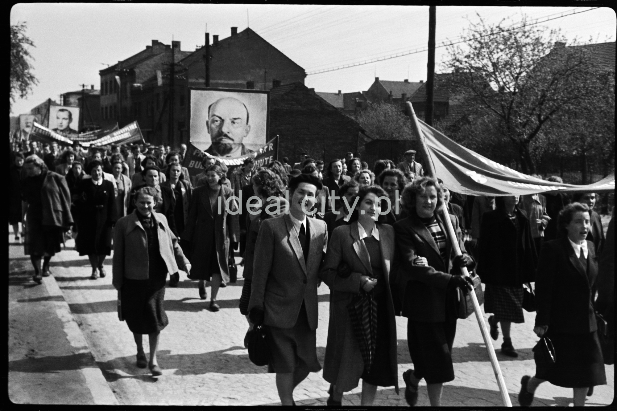 Ubrani odświętnie ludzie maszerują w pochodzie niosąc transparenty i portret Lenina.
