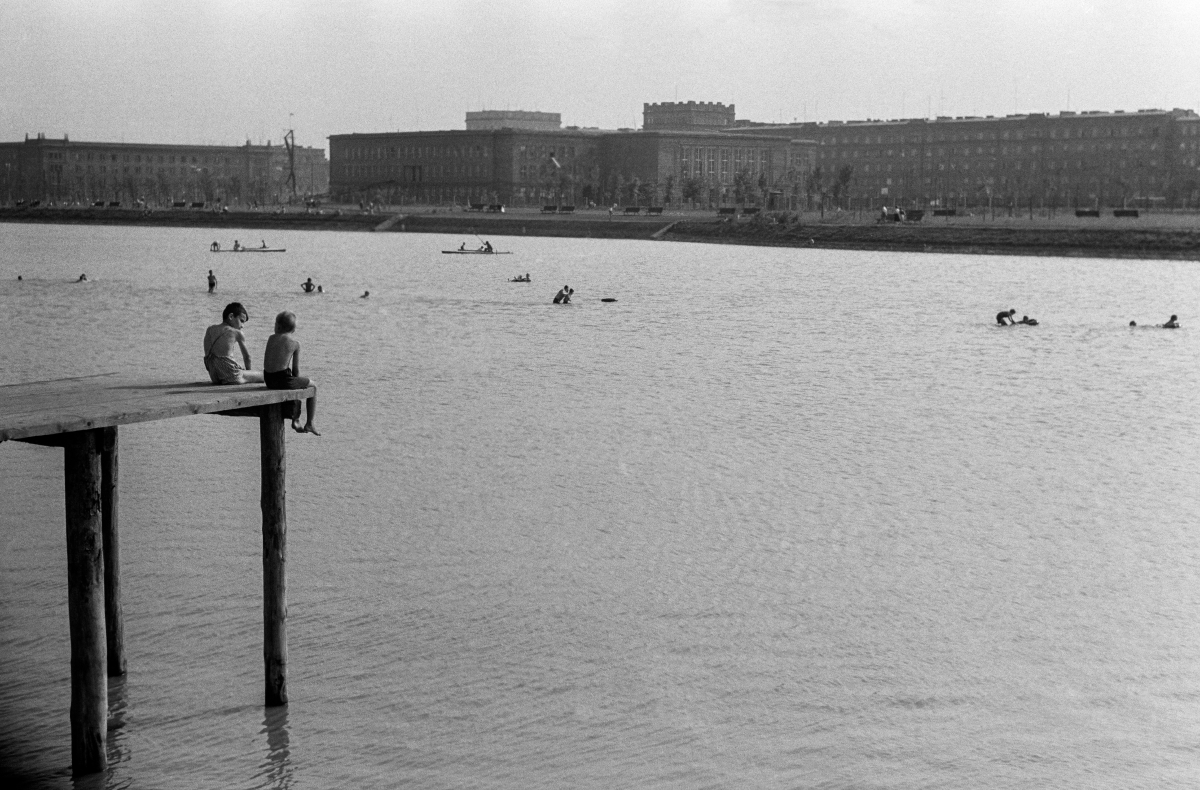 Two boys in bathing shorts are sitting on a high wooden platform overlooking the surface of the lagoon, and monumental city buildings on the other side of the river in the background.