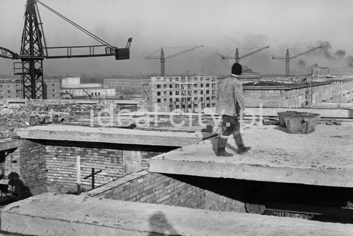 A laborer walks on the storey under construction, other buildings and construction cranes in the background.