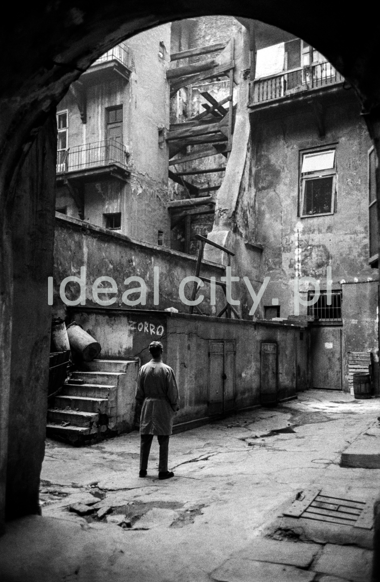 The man in the coat looks up the run-down yard of an old tenement house.