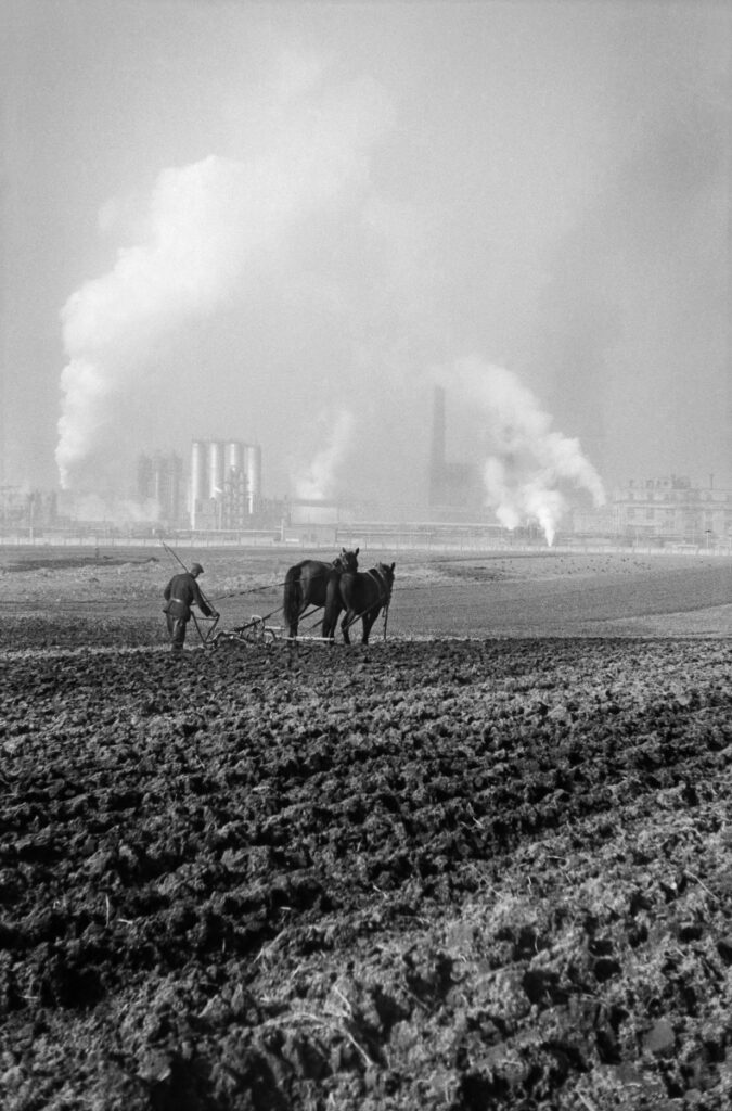 A farmer plows a field with a horse-drawn plow, in the background the smoking chimneys of the plant.