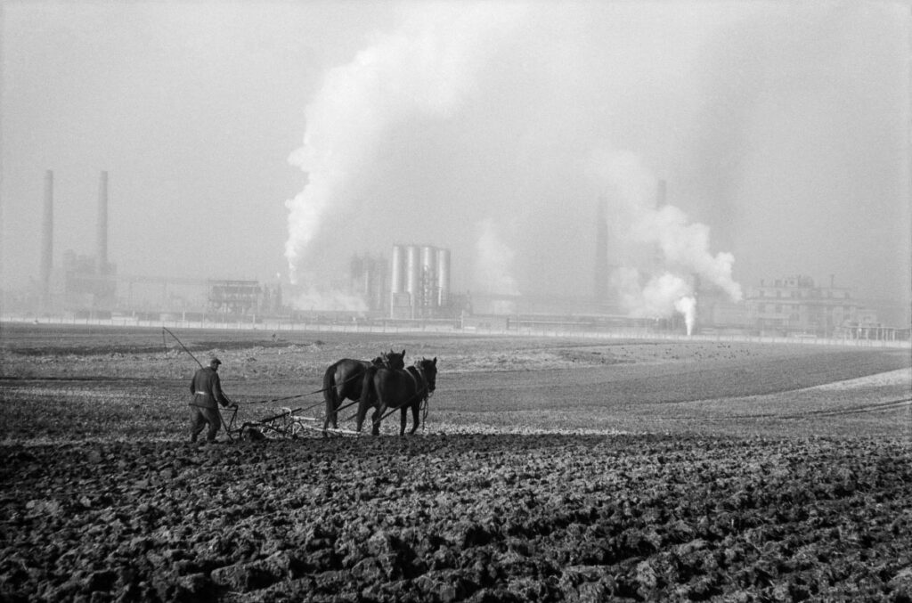 A farmer plows a field with a horse-drawn plow, in the background the smoking chimneys of the plant.