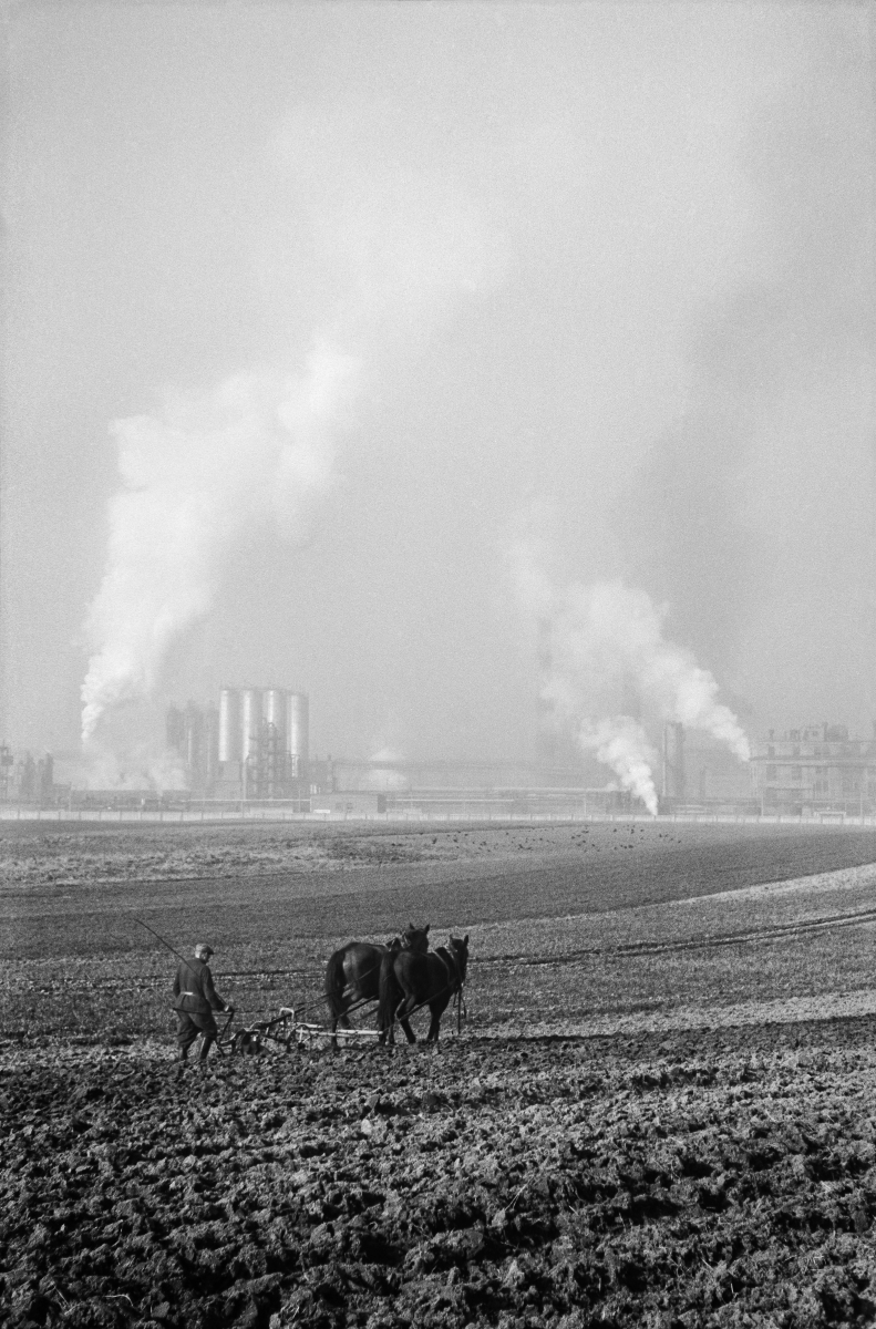 A farmer plows a field with a horse-drawn plow, in the background the smoking chimneys of the plant.