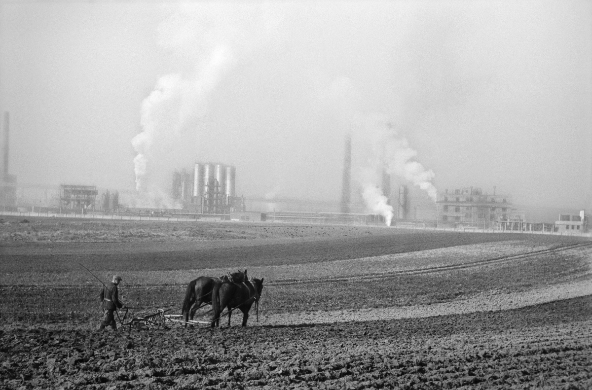 A farmer plows a field with a horse-drawn plow, in the background the smoking chimneys of the plant.