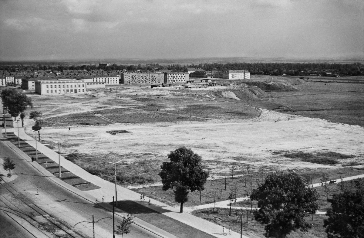 A view from a height on a trampled square, next to which a wide avenue with a tram line runs, in the background a block of residential buildings