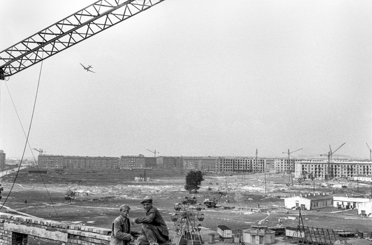 A wide perspective from a height over the city under construction, in the foreground two little figures of workers, an airplane in the sky.