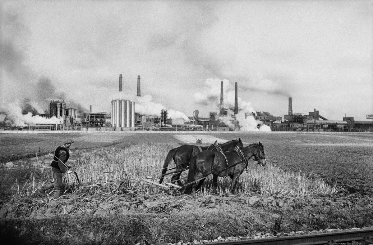 A farmer plows a field with a horse-drawn plow, in the background the smoking chimneys of the plant.