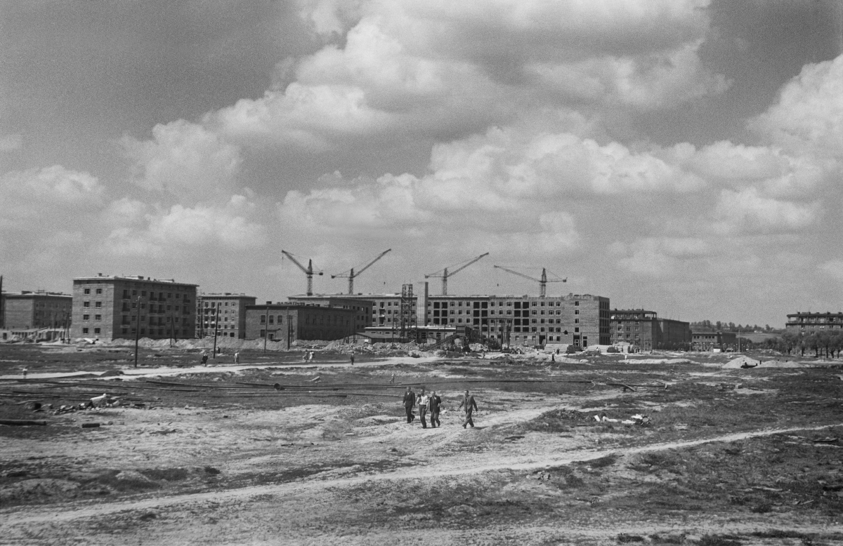 A view of the construction site, residential buildings in the background, in the center of the frame, a few small figures walk forward.