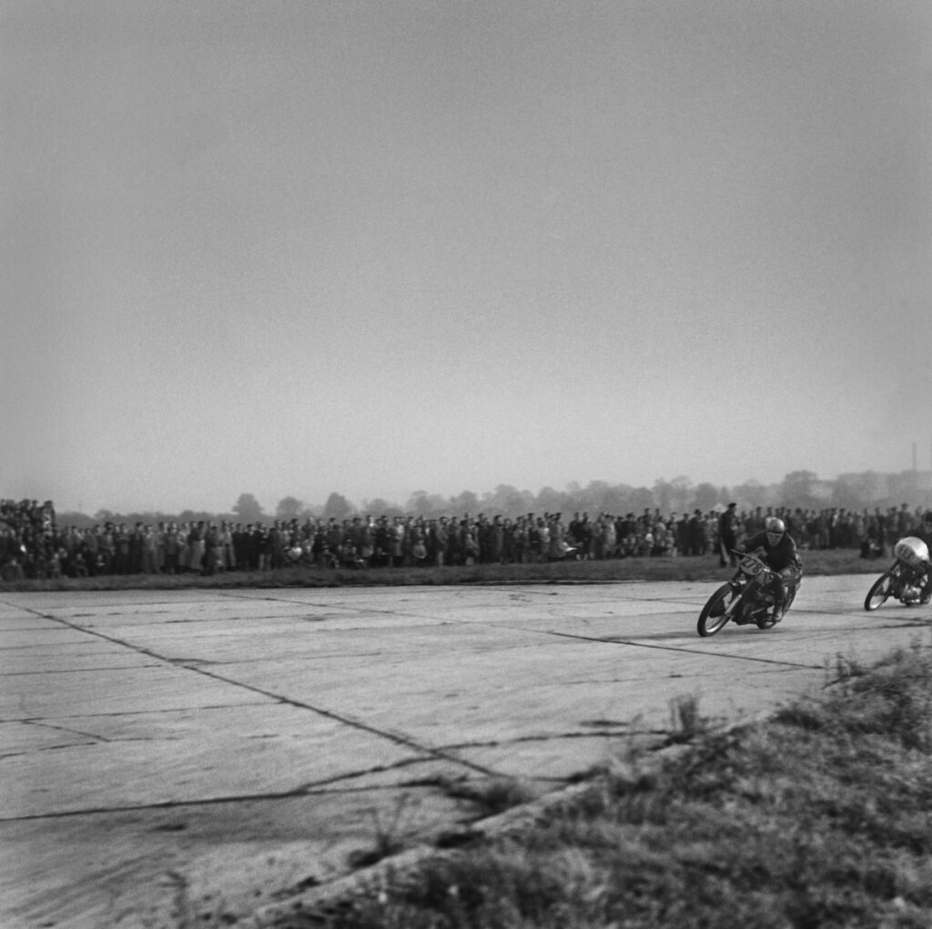 The motorcyclist is driving the bend at high speed, with spectators in the background.