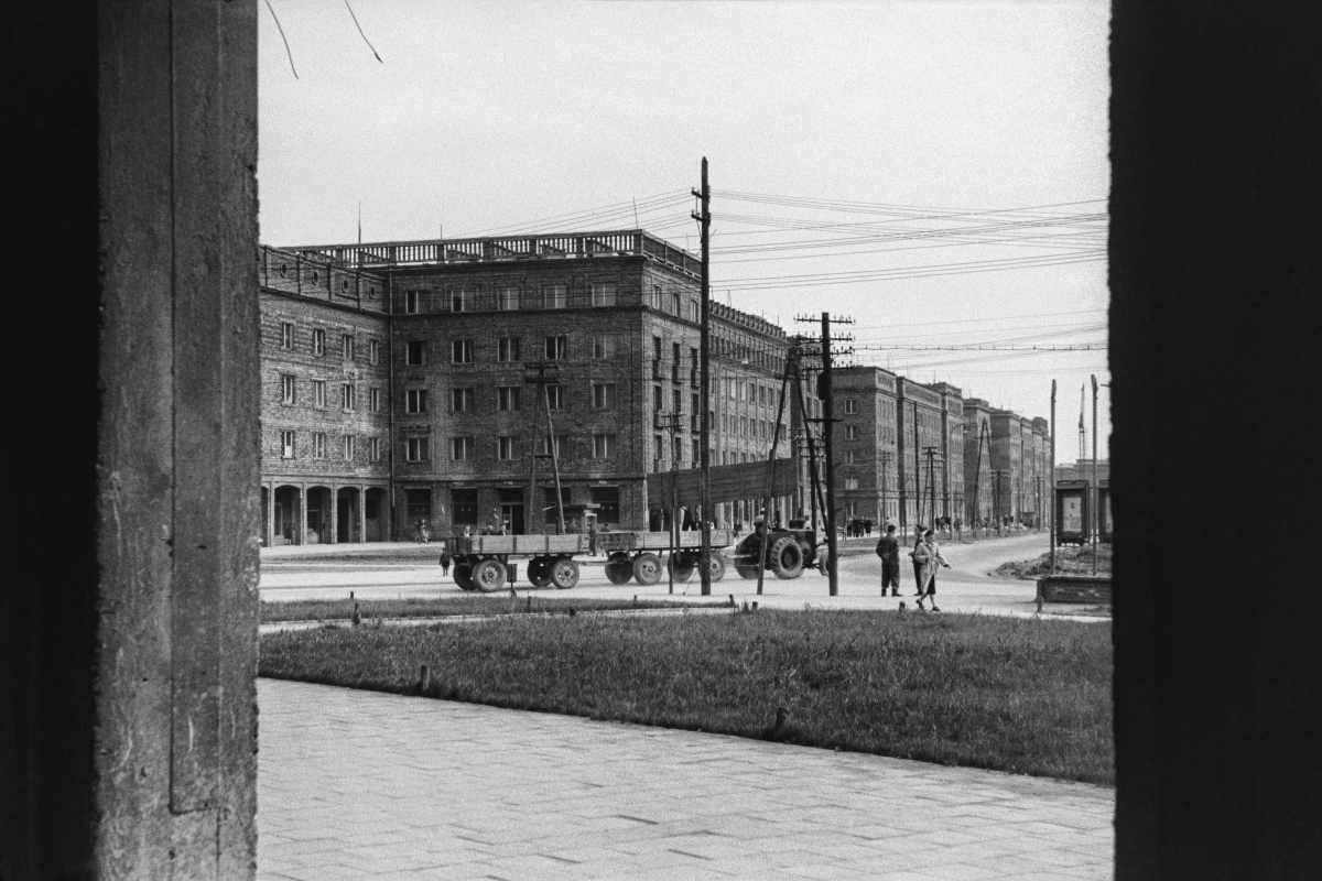 A view of a low, compact block of brick buildings, a tractor with trailers passing in the center of the frame, people walking by.