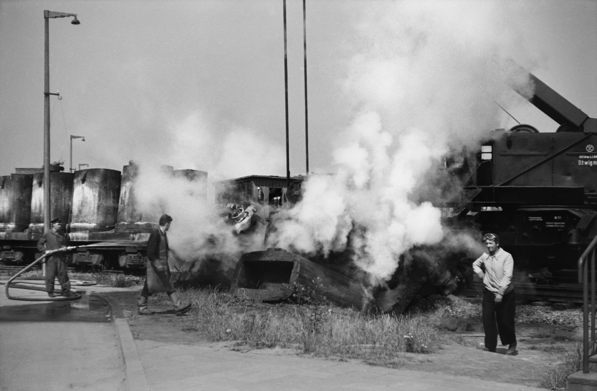 Steam puffs from extinguished water from the jets of objects lying next to the railway depot, near three men, one of them smokes a cigarette.