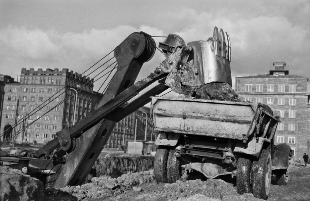 The excavator loads soil from the excavation onto a truck semi-trailer, residential buildings in the background.