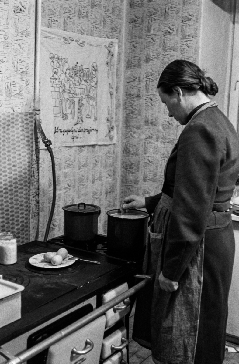 A woman with a scarf on her head and a kitchen apron looks after a meal that is cooking on the stove.