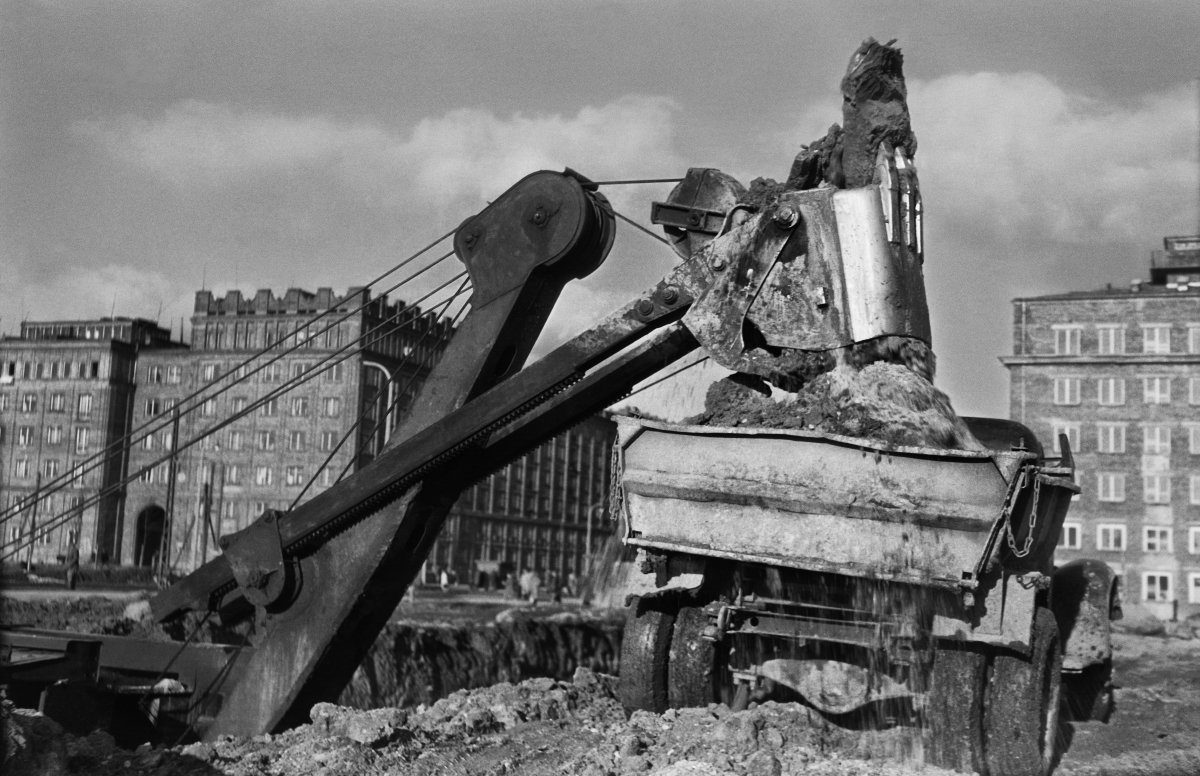 The excavator loads soil from the excavation onto a truck semi-trailer, residential buildings in the background.