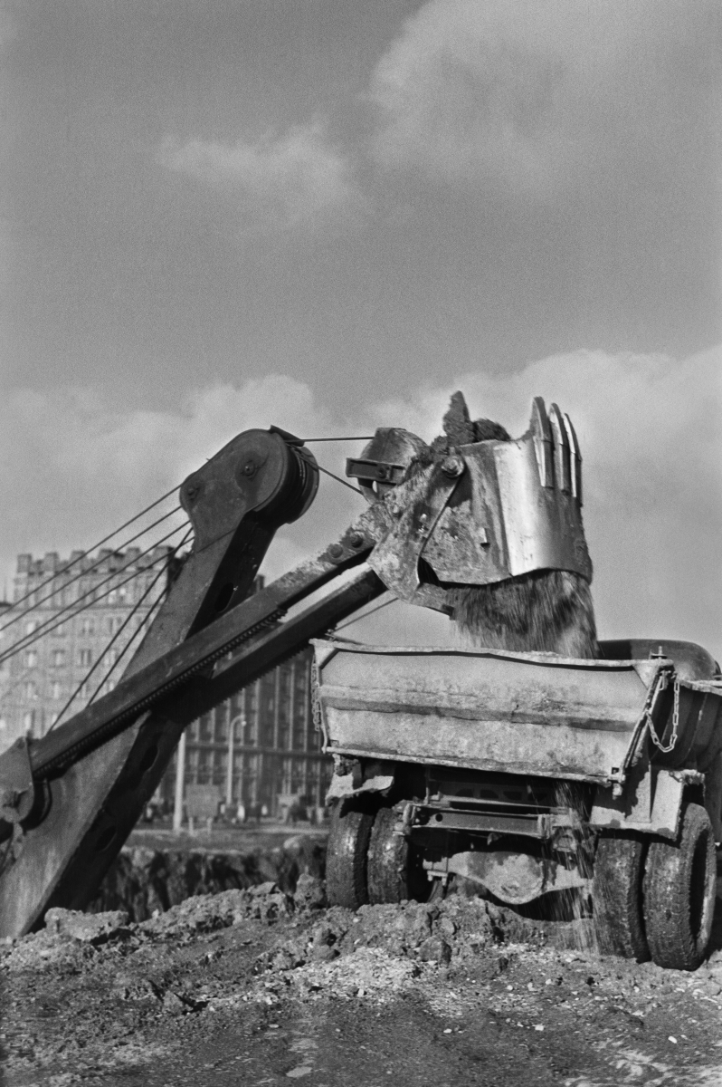 The excavator loads soil from the excavation onto a truck semi-trailer, residential buildings in the background.