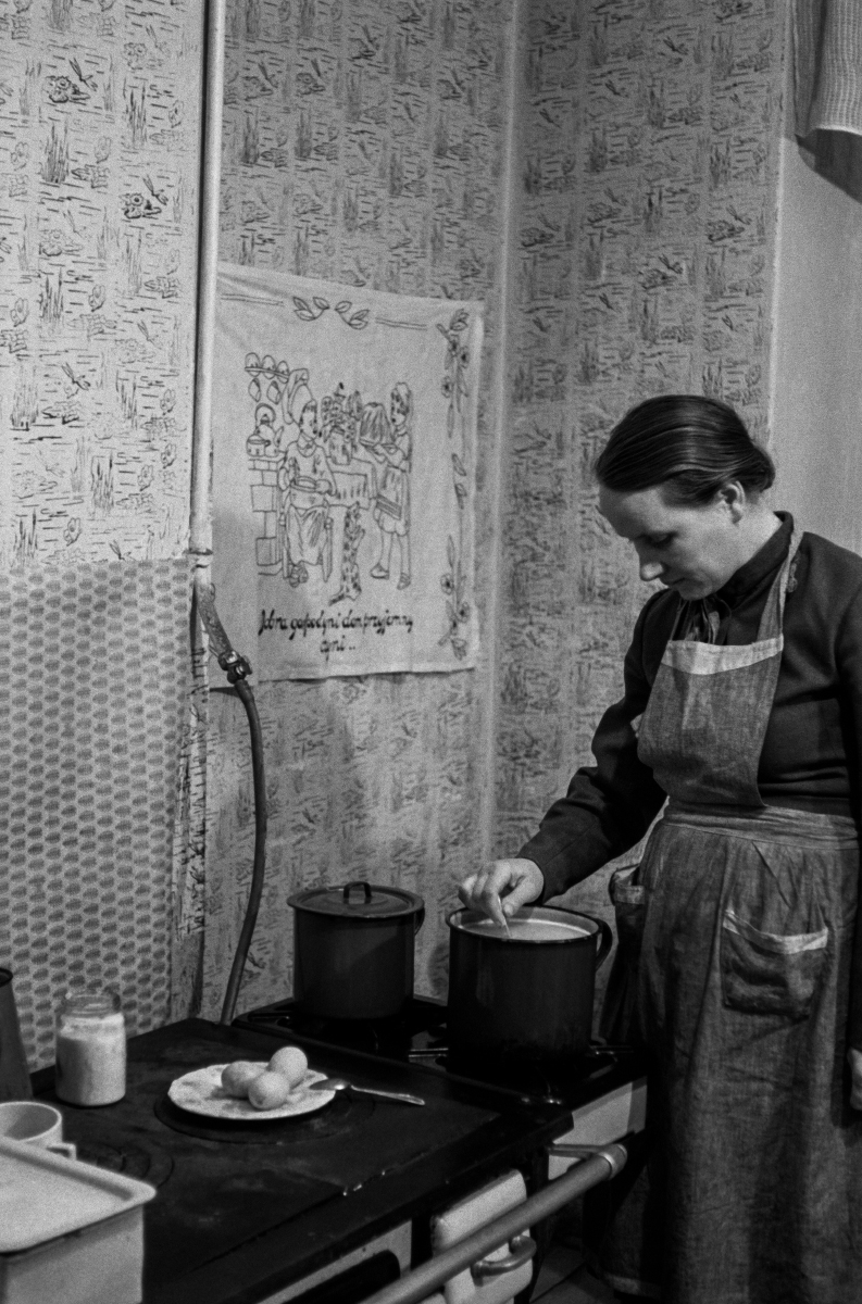 A woman with a scarf on her head and a kitchen apron looks after a meal that is cooking on the stove.