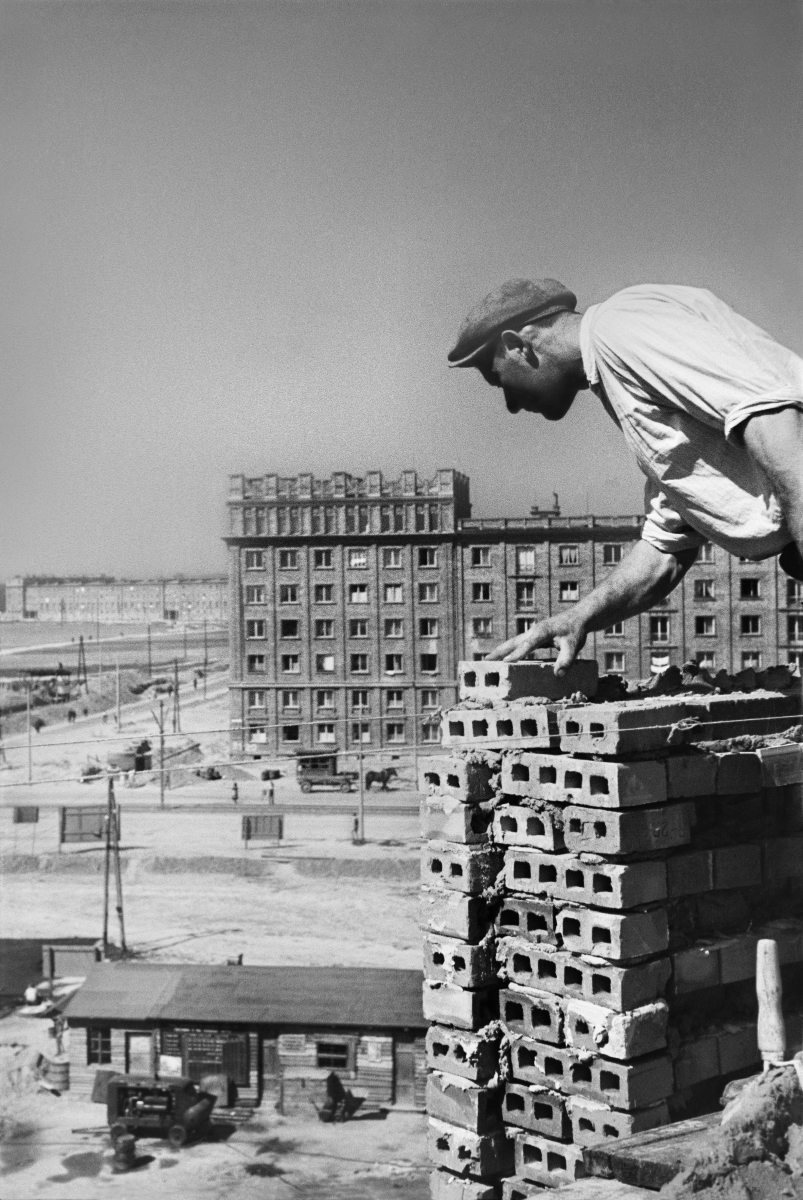 A bricklayer in a white shirt and flat cap places the bricks of the building on the upper storey, with a completed block in perspective.
