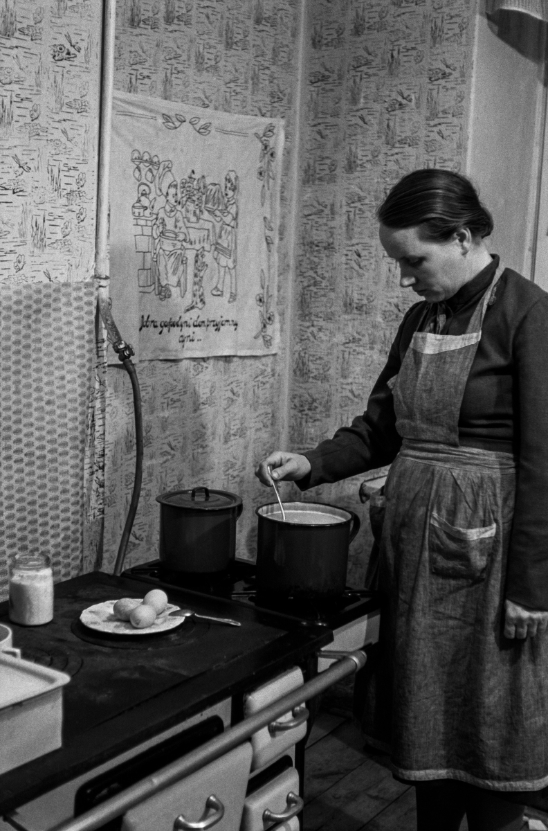 A woman with a scarf on her head and a kitchen apron looks after a meal that is cooking on the stove.