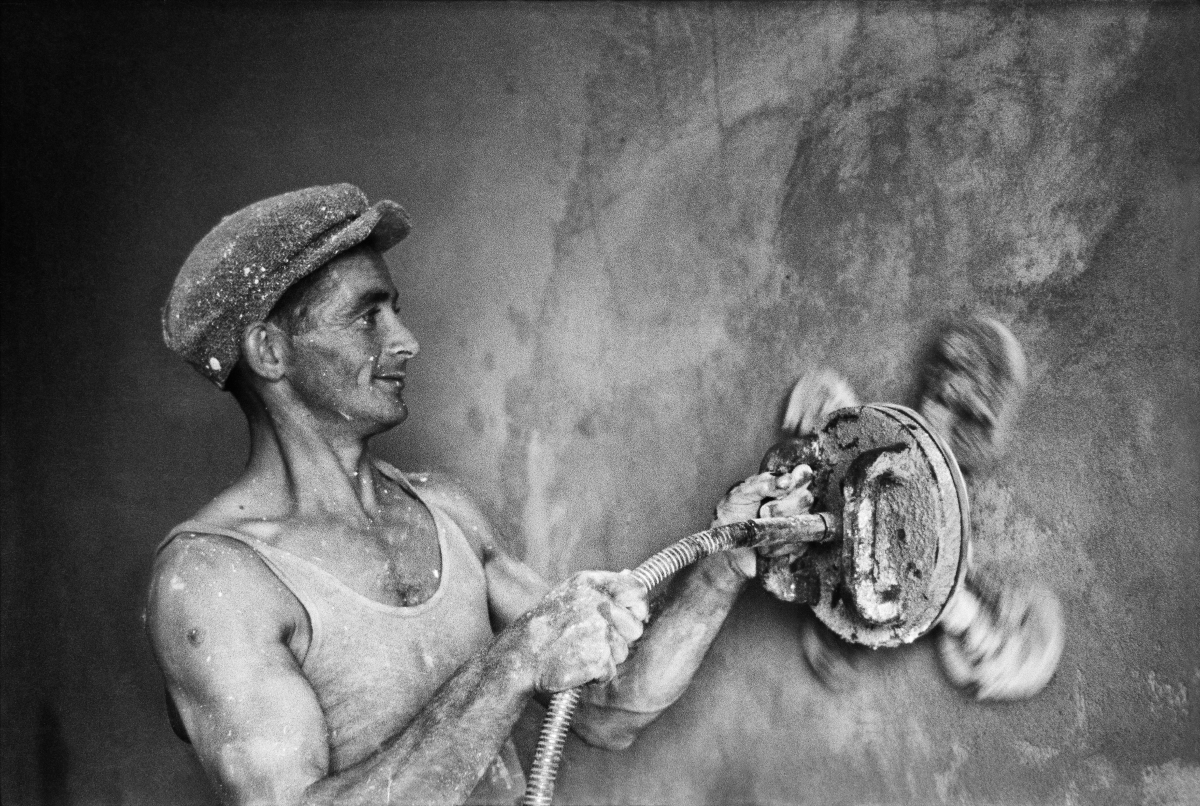 A worker in a tank top and a flat cap polishes the wall with an automatic grinder.