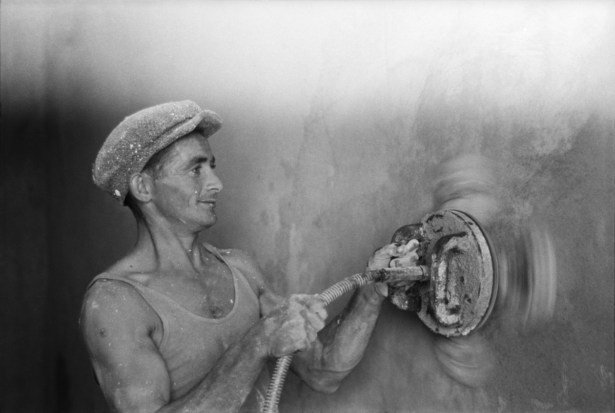 A worker in a tank top and a flat cap polishes the wall with an automatic grinder.