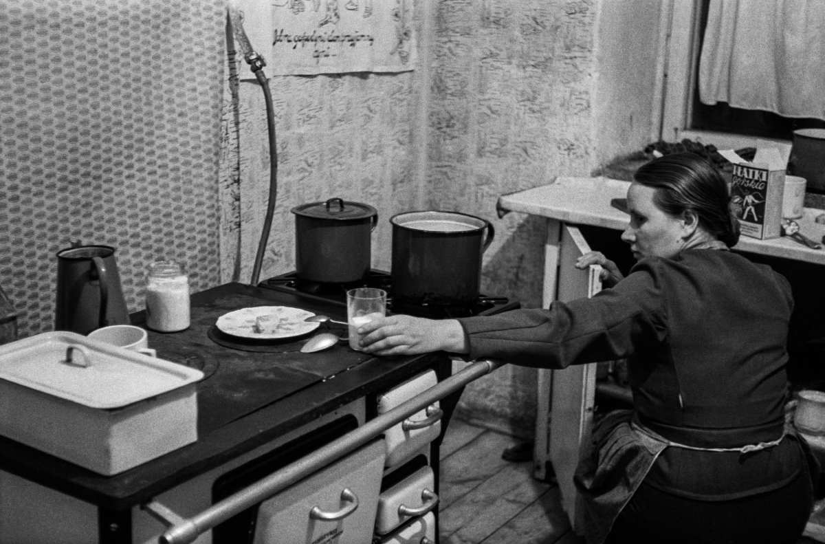 A woman with a scarf on her head and a kitchen apron looks after a meal that is cooking on the stove.