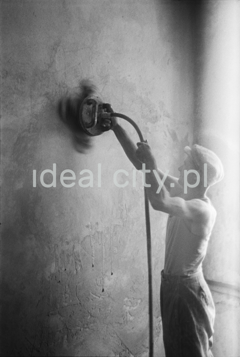 A worker in a tank top and a flat cap polishes the wall with an automatic grinder.