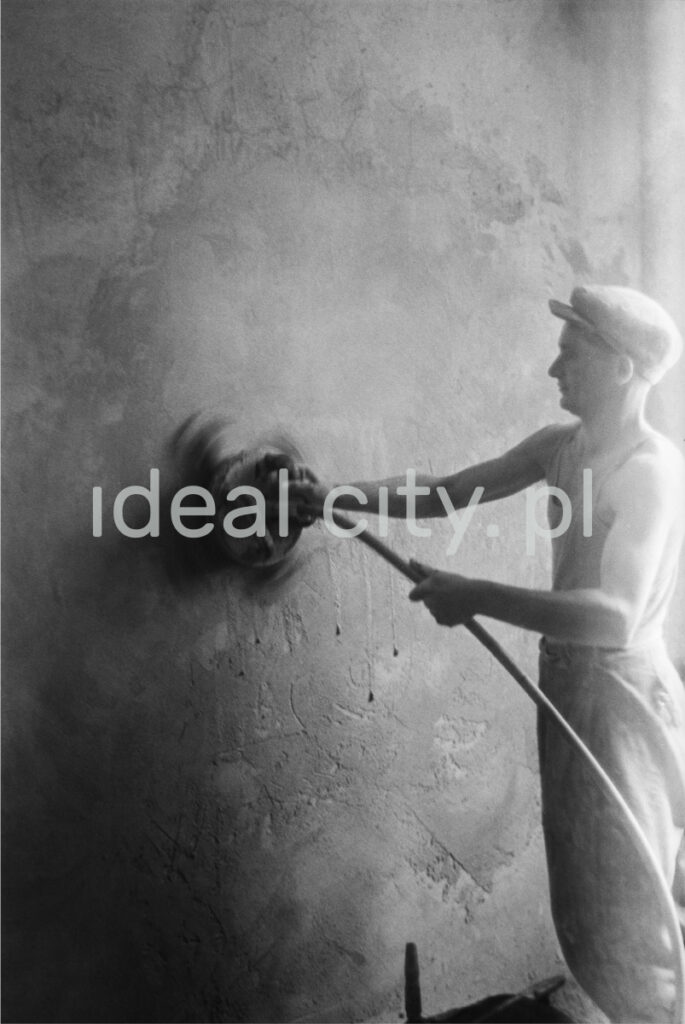 A worker in a tank top and a flat cap polishes the wall with an automatic grinder.
