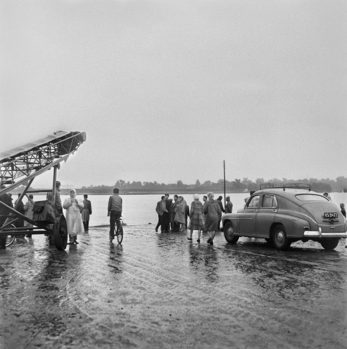 Onlookers gathered on the road that runs along the river bank. On the right, a parked car.
