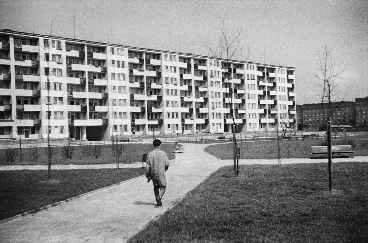 A man in a coat marches down the sidewalk toward a wide seven-story block.