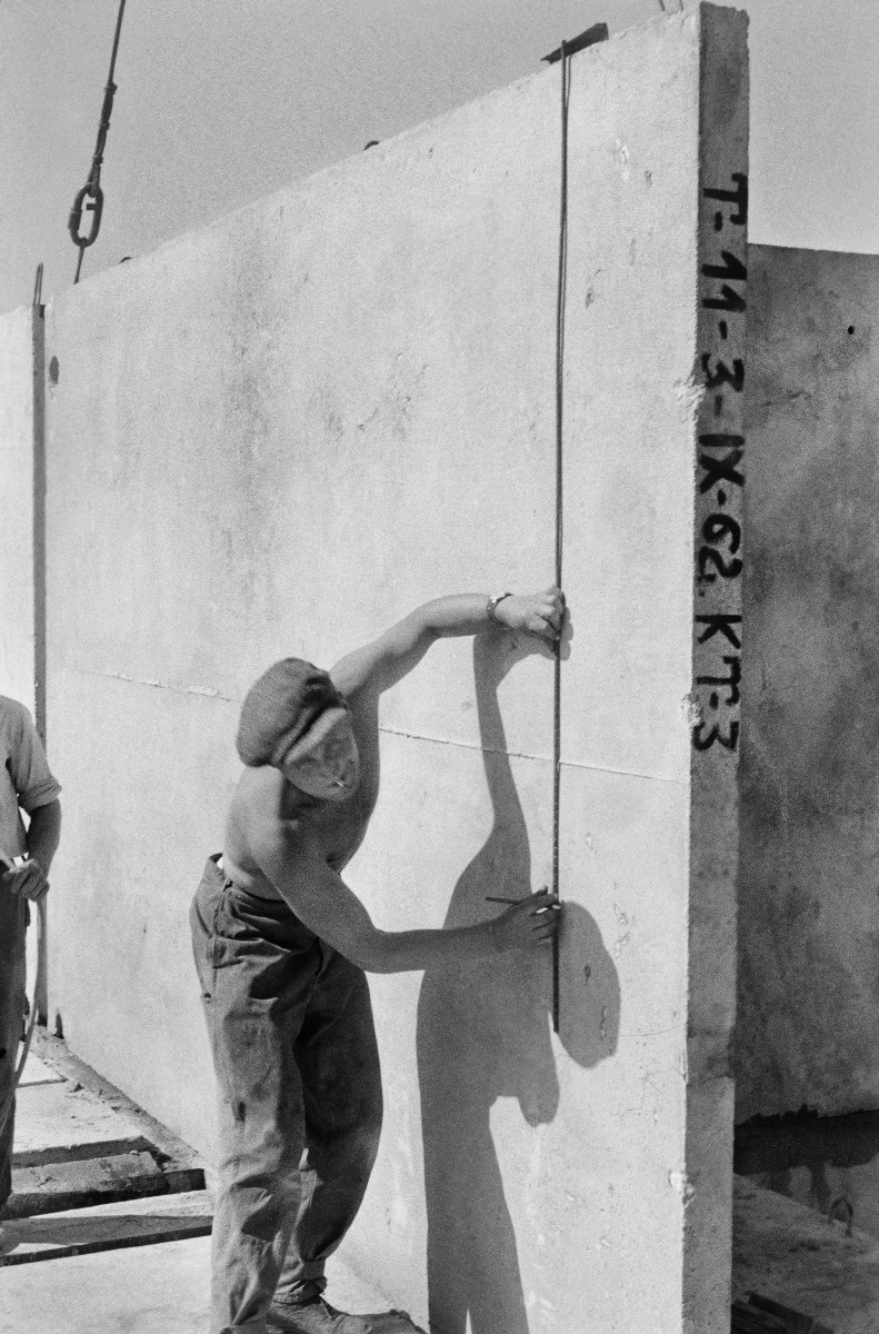 A shirtless worker in a flat cap places a prefabricated slab fed to him by a crane on the building's storey