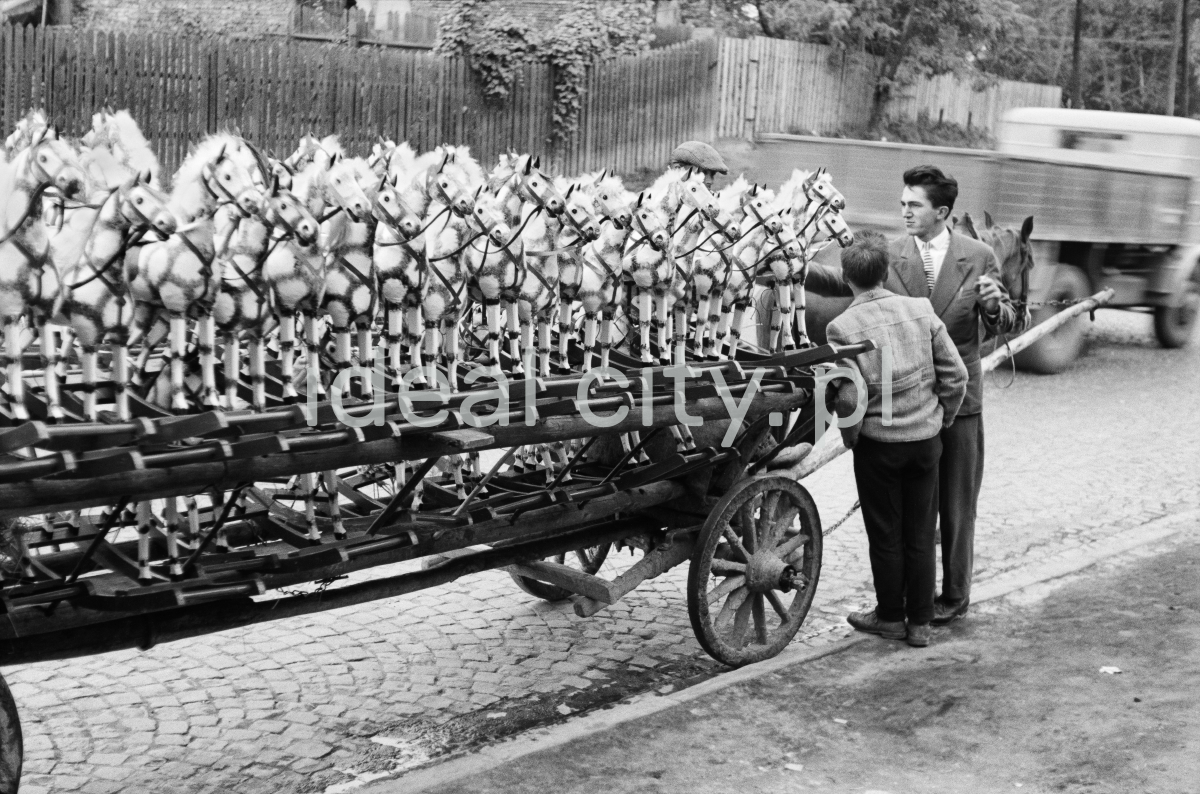 The shot shows a row of wooden, white-painted rocking horses placed on a wooden horse-drawn cart. On the right, an agent standing next to the workhorse is talking to a passerby.