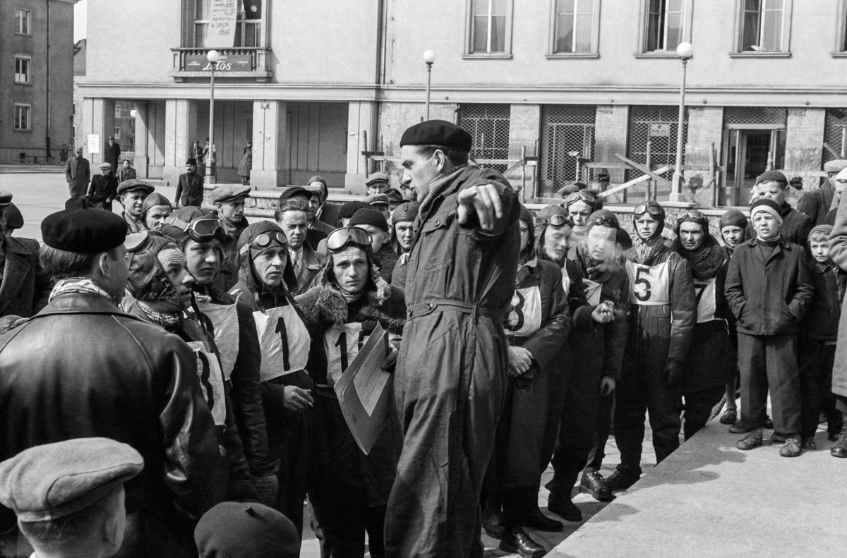 A man in a jumpsuit, standing on a platform, explains something, gesturing to the players gathered in front of him.