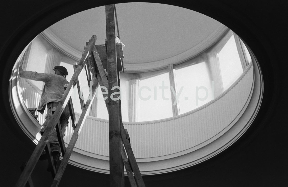 A man in work clothes, standing on a ladder, performs finishing works inside a circular skylight located in the ceiling of the building.