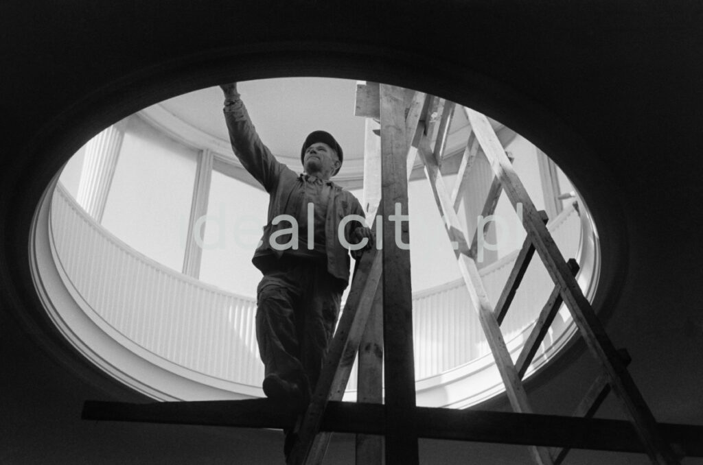 A man in work clothes, standing on a ladder, performs finishing works inside a circular skylight located in the ceiling of the building.