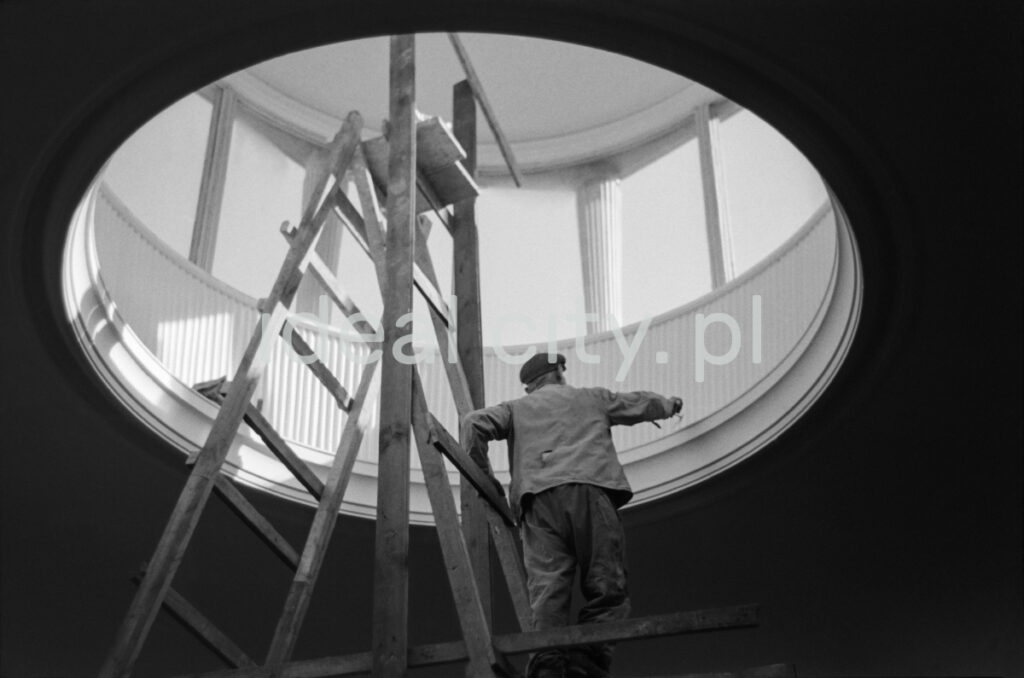 A man in work clothes, standing on a ladder, performs finishing works inside a circular skylight located in the ceiling of the building.