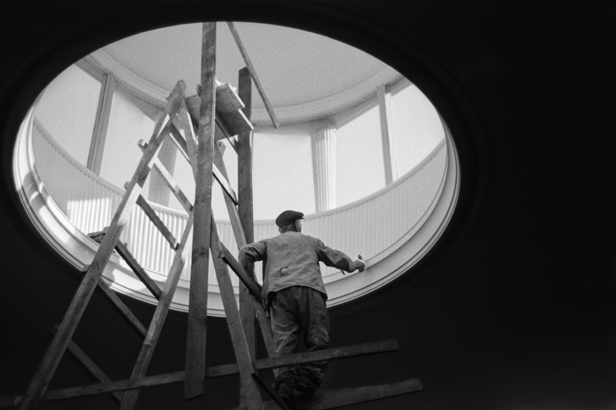A man in work clothes, standing on a ladder, performs finishing works inside a circular skylight located in the ceiling of the building.