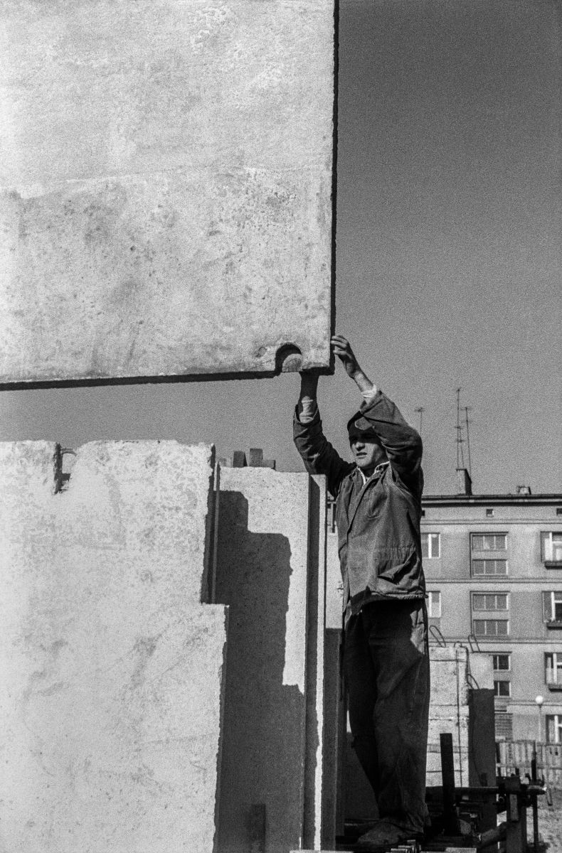 A man in work clothes steers a concrete slab fed by a crane.