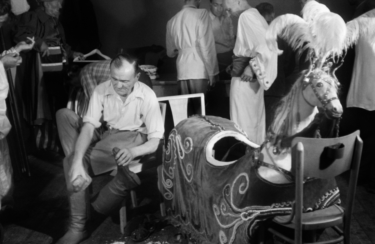 A man in a shirt, while sitting, wears boots with high uppers, next to a horse outfit with a headpiece.
