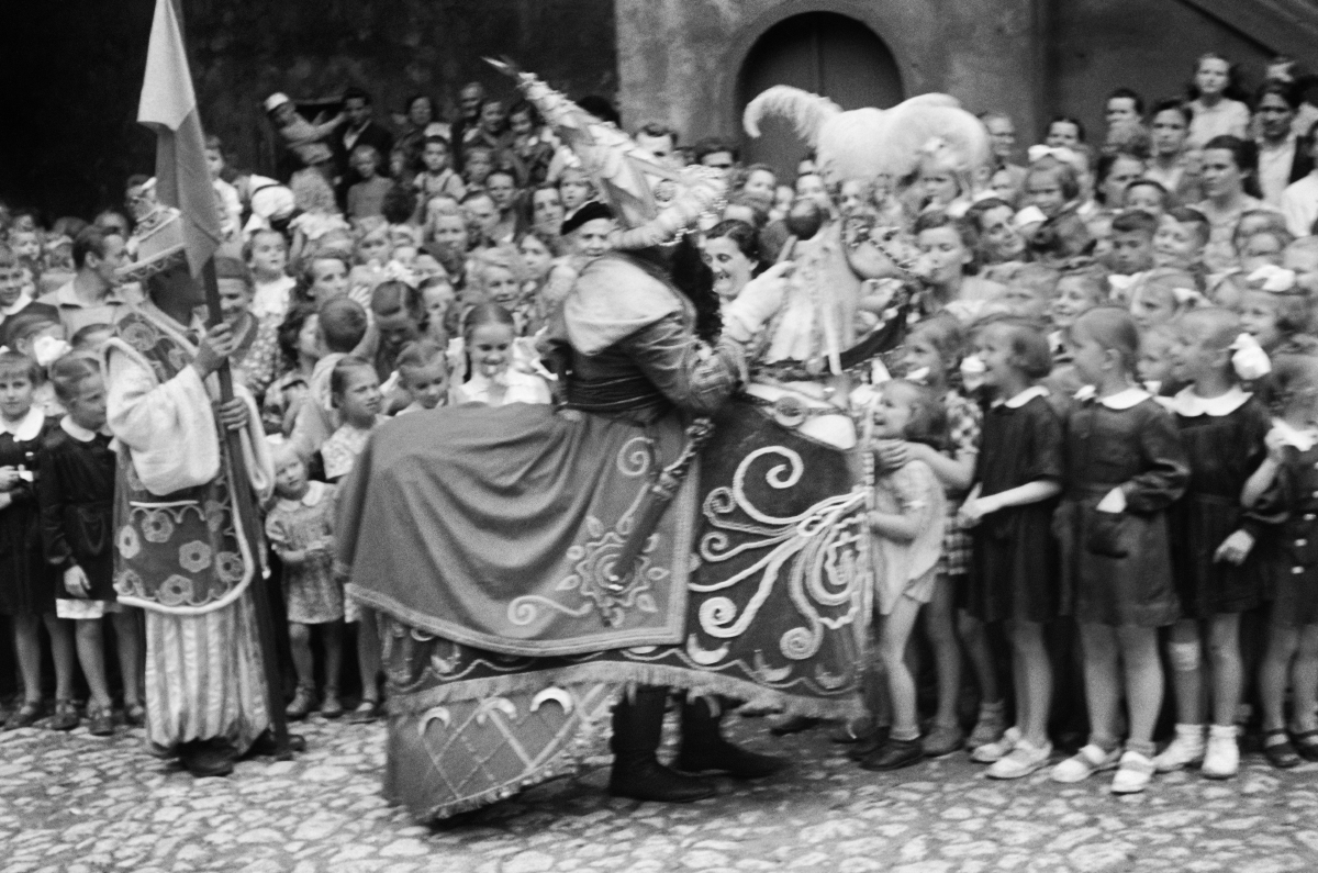 A man dressed as a horse with a purse on his head walks alongside the crowd on the street.