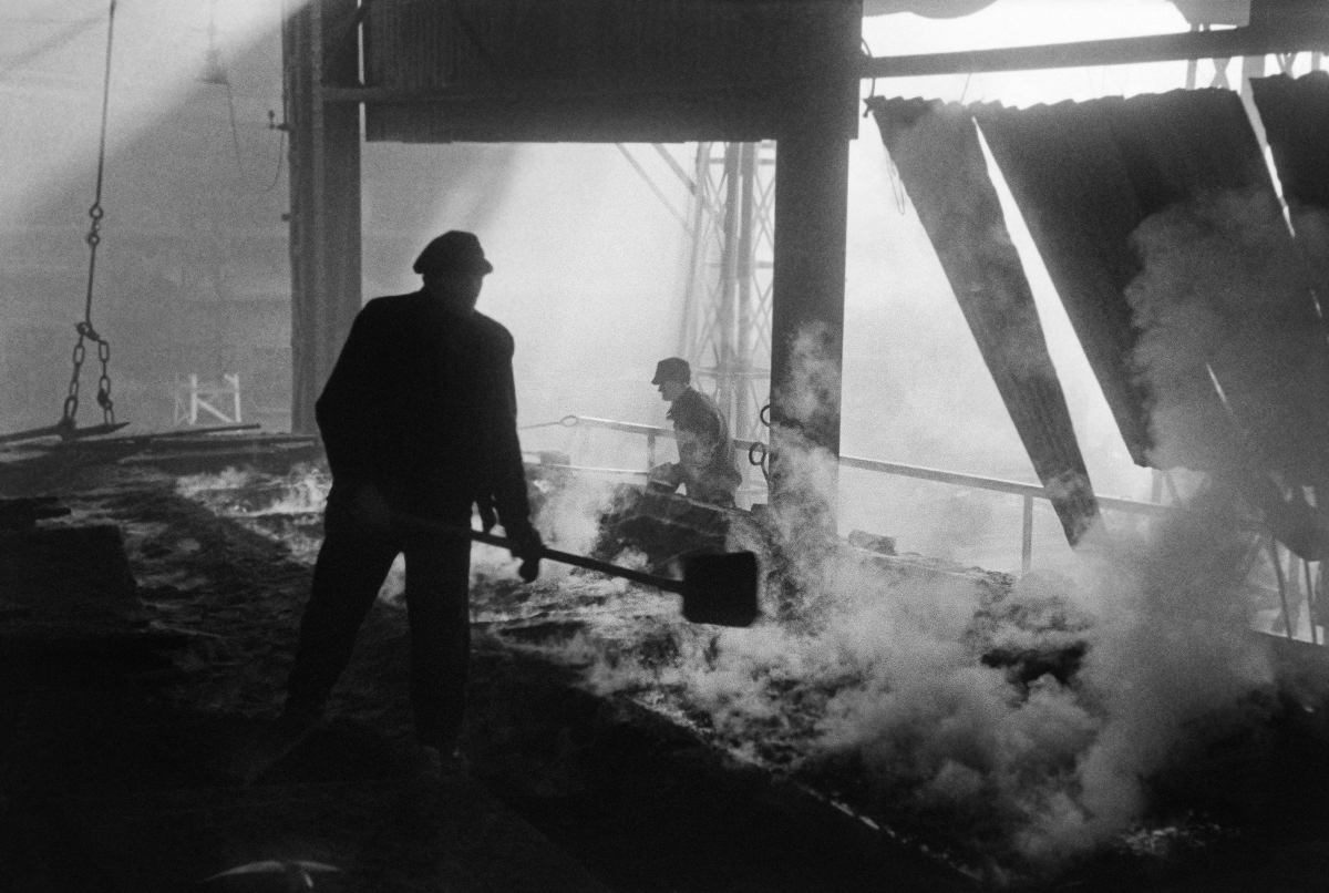 Two men in work clothes are operating shovels over the smoking substance in the production hall.