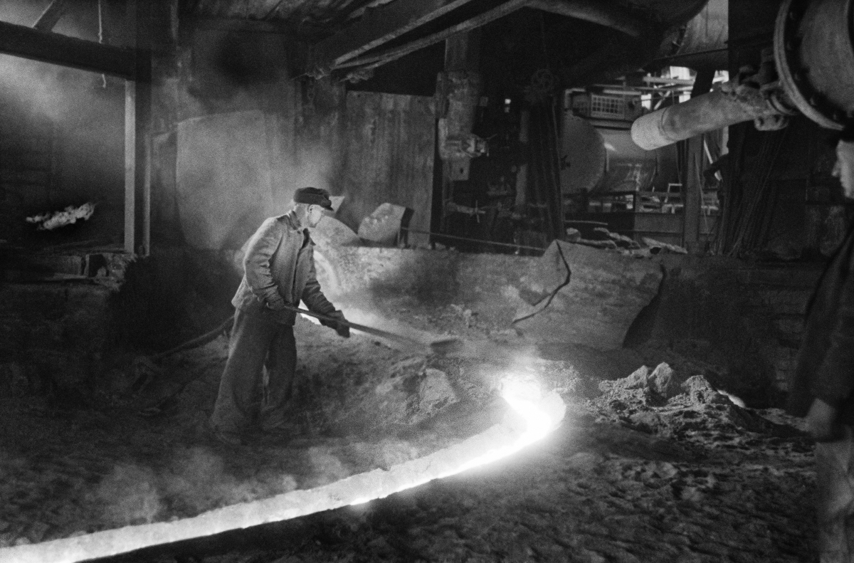 A man in work clothes uses a shovel to correct a stream of flowing iron.