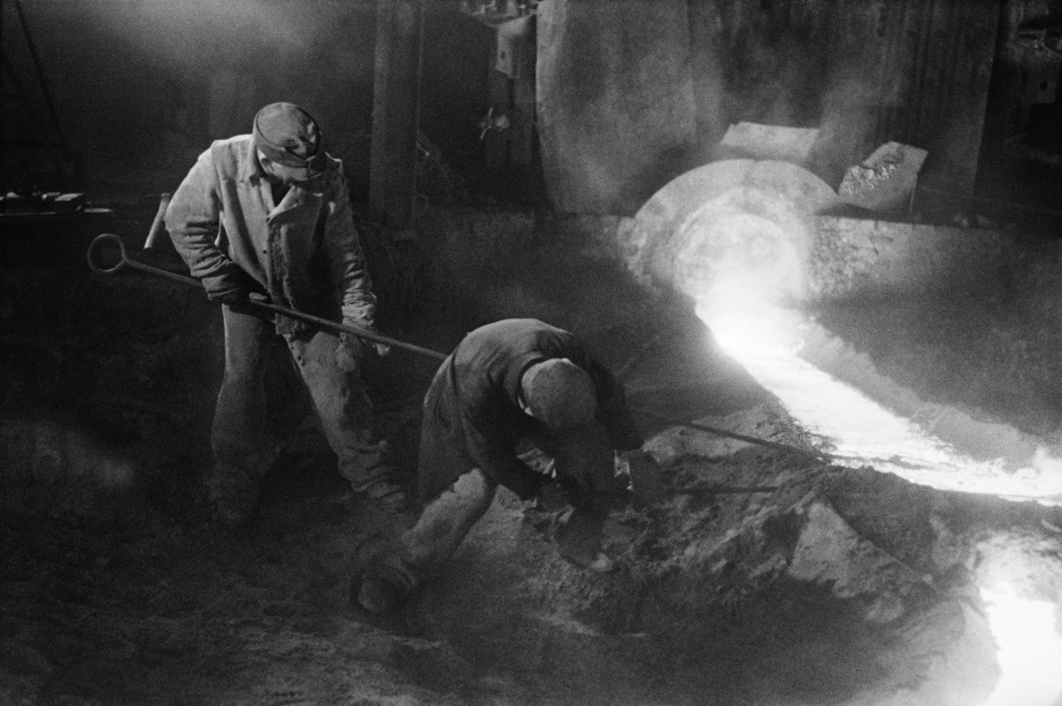 Two men in working clothes use long rods to correct a stream of flowing iron.
