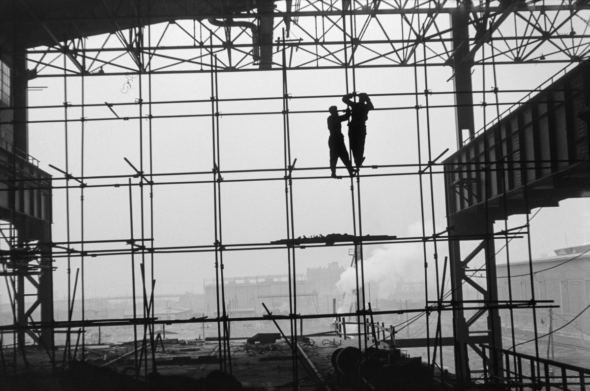 Silhouettes of two men in work clothes on scaffolding along the wall of a newly built hall, factory landscape in the background.