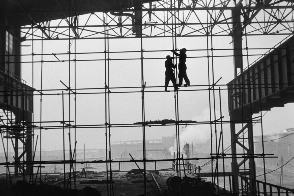 Silhouettes of two men in work clothes on scaffolding along the wall of a newly built hall, factory landscape in the background.