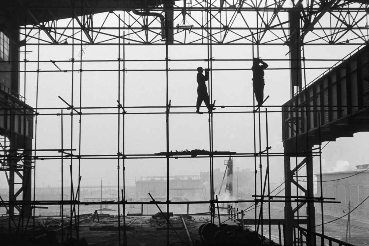Silhouettes of two men in work clothes on scaffolding along the wall of a newly built hall, factory landscape in the background.