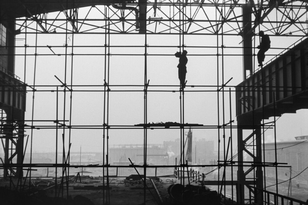 Silhouettes of two men in work clothes on scaffolding along the wall of a newly built hall, factory landscape in the background.