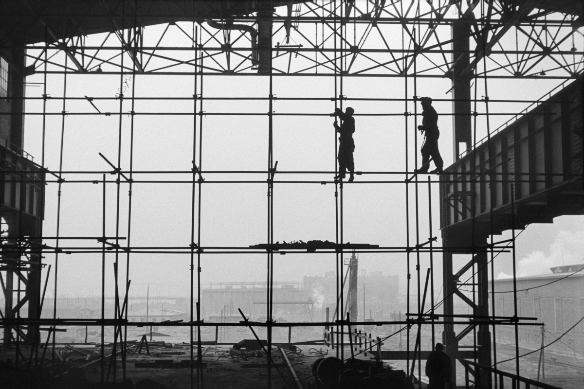 Silhouettes of two men in work clothes on scaffolding along the wall of a newly built hall, factory landscape in the background.