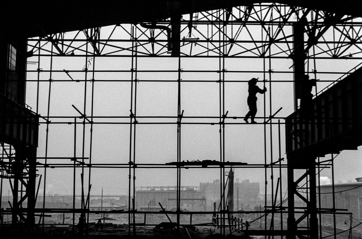 Silhouettes of two men in work clothes on scaffolding along the wall of a newly built hall, factory landscape in the background.