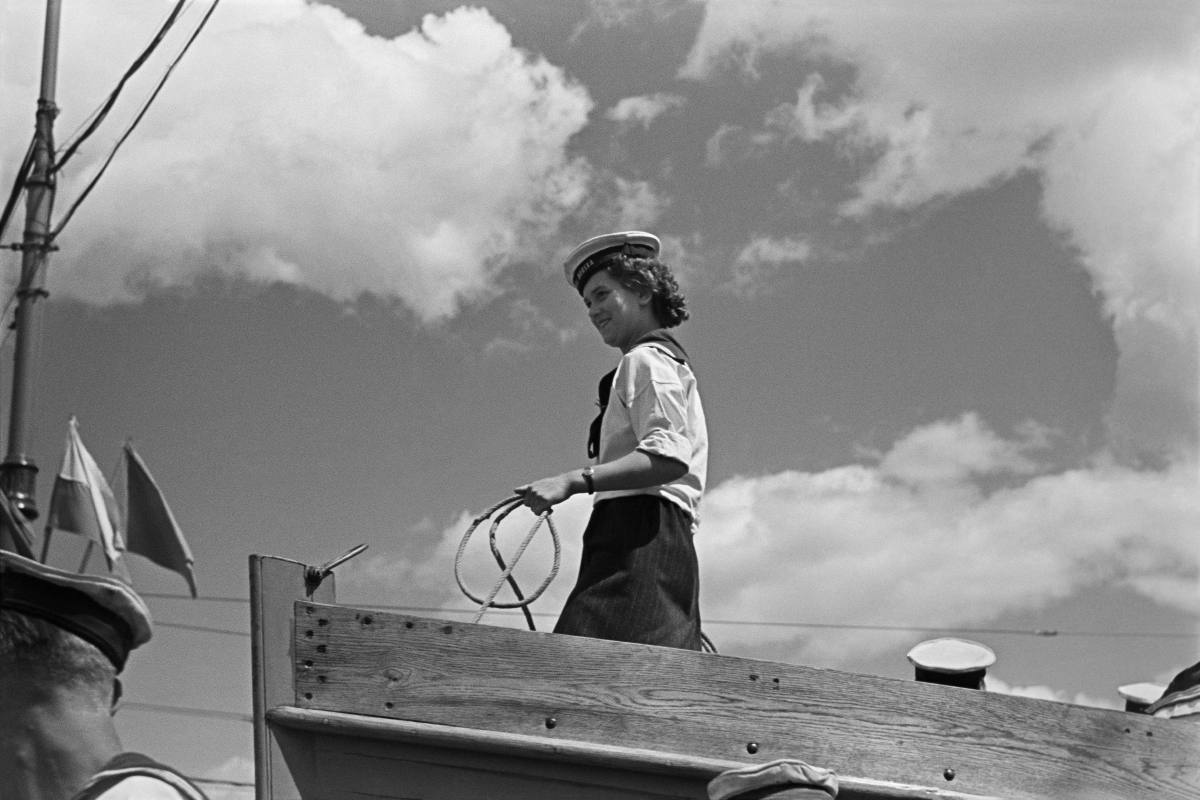 A young woman in a sailor suit stands at the front of the boat holding a rope in her hand.