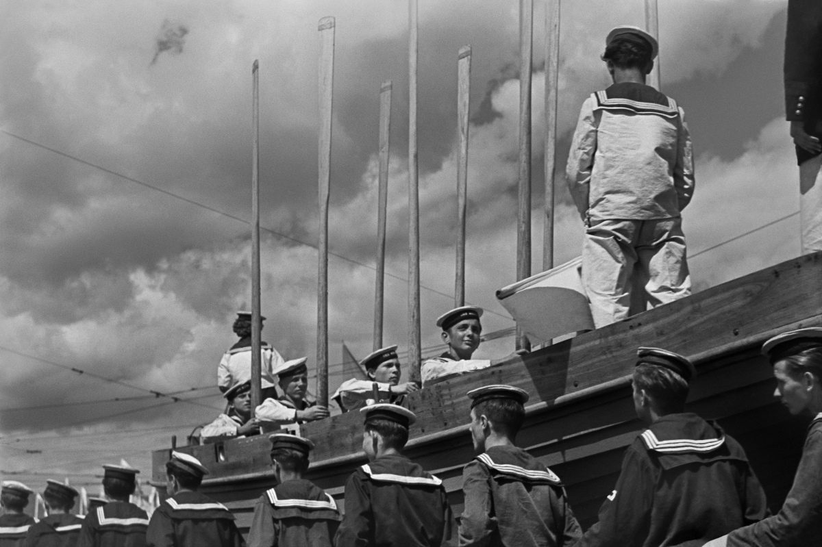 Young men in sailor's outfits stand with their oars upright in a wooden boat placed on a raised platform, below are others marching along the side.